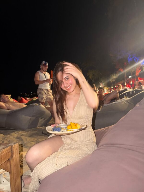 Jannat Zubair smiling at a beachside lounge at night while holding a dessert plate, wearing a light beige outfit under warm ambient lights.